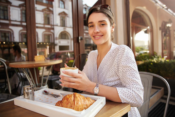 Photo of attractive young brunette woman with sunglasses on her head having breakfast over summer terrace, keeping cup of coffee in raised hands and smiling widely to camera