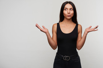 Suprised young attractive long haired brunette female rounding eyes while looking positively aside and raising palms up, dressed in casual black clothes while posing over white background