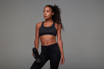 Studio shot of young slim dark skinned curly brunette woman in athletic black top and leggins, looking thoughtfully aside and keeping fitness bottle in hand, isolated over grey background