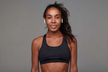 Obraz premium Close-up of young pretty brown-eyed dark skinned brunette woman wearing earpones in her ears and looking positively at camera, isolated over grey background
