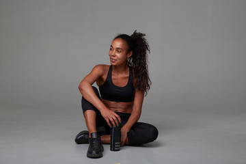Studio photo of long haired dark skinned curly lady in sporty black wear relaxing after hard training, sitting on floor with fitness bottle, isolated over grey background