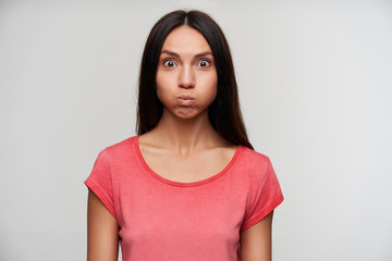 Obraz premium Indoor shot of open-eyed young brunette lady with natural makeup puffing out cheeks while looking surprisedly to camera, posing over white background with hands down