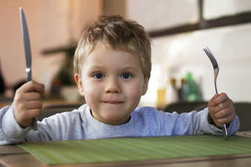 A little smiling boy with cutlery in his hands, sits at a table in a cafe and waits for his...