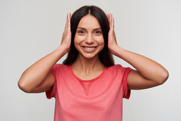 Cheerful young lovely dark haired lady with natural makeup closing her ears with hands and looking happily at camera and smiling widely, standing against white background