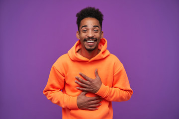 Portrait of happy lovely young curly dark skinned brunette male with beard keeping raised palms on his chest while rejoicing about good news and smiling joyfully, isolated over purple background