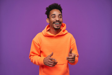 Cheerful young pretty bearded dark skinned man with curly black hair laughing happily and showing aside with raised forefingers, standing against purple background in casual sporty wear