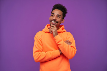 Indoor shot of positive young brown-eyed bearded brunette male with dark skin holding his chin and smiling dreamily while posing over purple background in sporty casual wear