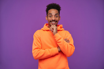 Cheerful young pretty brown-eyed dark skinned brunette man with beard holding raised hand on his chin and smiling happily to camera, isolated over purple background in orange hoodie