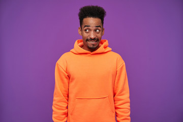 Studio shot of young lovely bearded dark skinned curly brunette guy looking positively aside with confused grimace, keeping hands down while standing over purple background