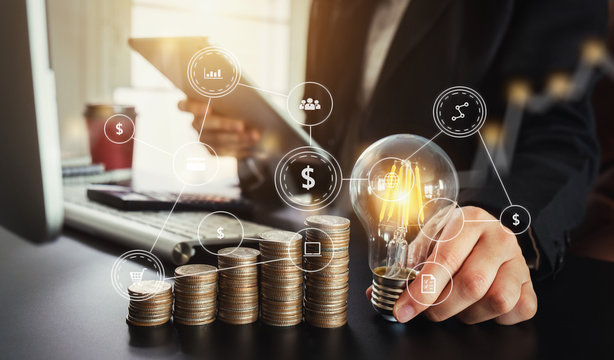 business man hand holding lightbulb with using smartphone and calculator to calculate and money stack. idea saving energy and accounting finance in morning light 