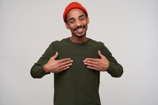 Pleased young dark haired bearded dark skinned guy holding palms on his chest and smiling happily to camera, being in nice mood while standing over white background