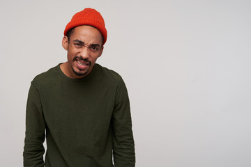 Indoor shot of young dark bearded dark skinned brunette guy looking to camera with dissatisfied gromace and frownig eyebrows, dressed in casual wear while standing over white background