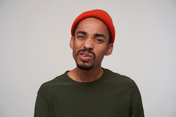 Displeased young pretty bearded dark skinned brunette guy twisting his mouth and looking at camera with pout, wearing red hat and khaki sweater while standing over white background