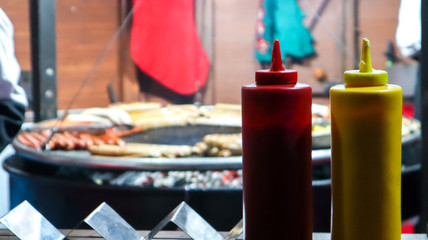 ketchup and mayonnaise in plastic tubes near a street grill in the foreground close-up with selective focus. Barbecue, street food festival. Two cans of meat sauces. Red and yellow container.
