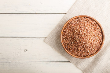 Wooden bowl with unpolished brown rice on a white wooden background. Top view, copy space.