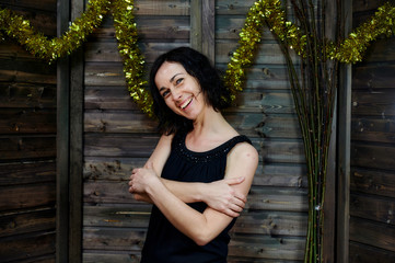 Portrait of a pretty smiling brunette woman in black dress on wooden background in vintage festive interior. Standing right in front of the camera with vivid emotions.