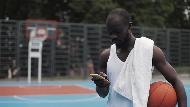 Young Muscly Afro - American Guy in White Singlet Holding Ball and Using his Smartphone Scrooling and Typing Message Walking at Street Basketbal Court. Technology and Sport Concept. Front View.