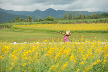 Fototapeta na wymiar asia woman with a hat in her hand walks in a field with field flowers and smiles sincerely, happy enjoying summer in yellow field at sunset. smiling with arms raised up. concept of freedom.