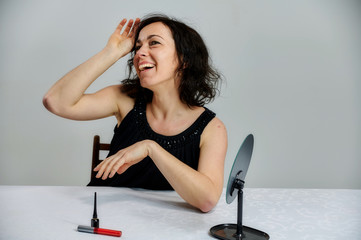 Portrait of a cute smiling talking brunette woman in a black dress on a white background. Sits at a table right in front of the camera with vivid emotions with a set of cosmetics.