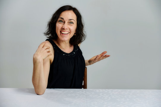 Model Sits at a table right in front of the camera with vivid emotions. Portrait of a cute smiling talking brunette woman in a black dress on a white background.