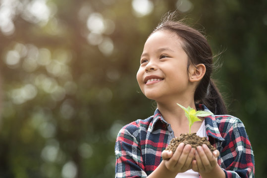 Kid Holding Young Plant In Hands Against Spring Green Background. Environment Earth Day In The Hands Of Trees Growing Seedlings. Concept Ecology