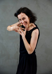 Fototapeta premium Model Standing right in front of the camera with vivid emotions. Vertical portrait of a cute smiling brunette woman in black dress on gray alternative background.
