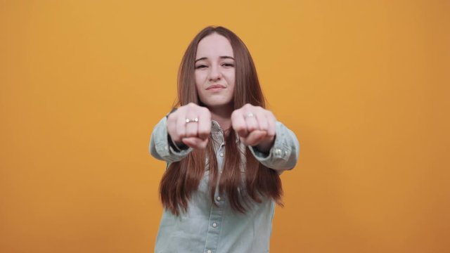 Caucasian attractive woman in denim blue shirt showing fists at camera,ready for fight isolated on orange background in studio. People emotions, lifestyle concept.