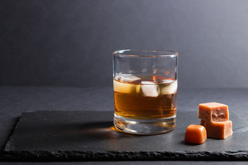 Glass of whiskey with ice and caramel candies on a black stone slate board on black background. Side view, selective focus, copy space.
