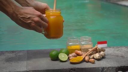 A hand of a man takes a glass with national balinese drink Jamu from the side of a swimming pool