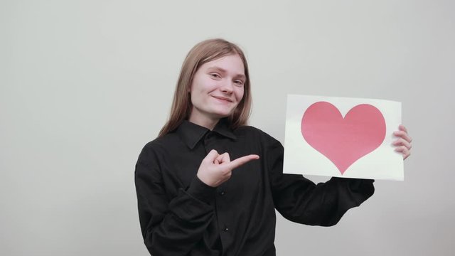 Charming caucasian young woman in fashion black shirt isolated on gray background in studio keeping paper with picture of red heart, pointing finger on it. People sincere emotions, lifestyle concept.