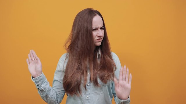 Disappointed attractive woman in denim blue shirt keeping crossed hands, doing stop, NO gesture isolated on orange background in studio. People emotions, lifestyle concept.