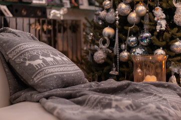 Christmas still life with grey nordic knit blanket and cushion with deers pattern on a couch, big candles on a floor and decorated Christmas tree on the back