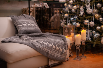 Christmas still life with grey nordic knit blanket and cushion with deers pattern on a couch, big candles on a floor and decorated Christmas tree on the back
