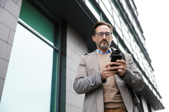 Image of mature bearded businessman using cellphone while walking