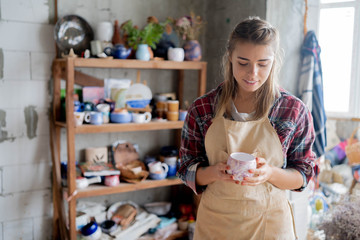 Young lovely smiling girl with blonde hair in apron holding a clay pot in her hands and standing in pottery studio. Pottery workshop. Clay model.