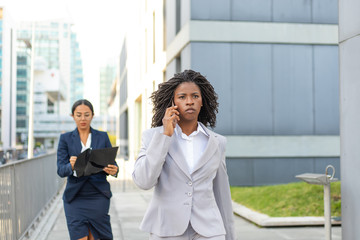 Fototapeta premium Focused young female professional talking on cell on her way to office. Young African American business woman walking outside in city. Communication concept