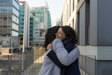 Happy excited multiethnic colleagues hugging each other near office building. Business women meeting outside. Old friends meeting at work concept