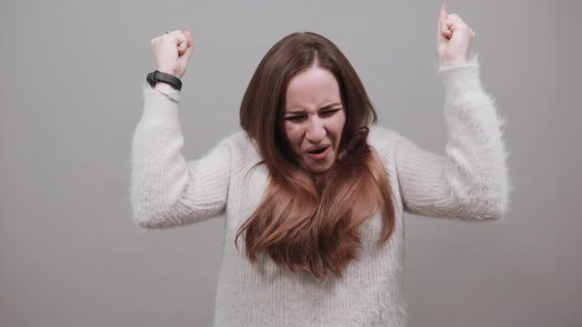 Cheerful charming caucasian woman wearing fashion blue sweater, dancing, smiling, keeping fist up isolated on gray background in studio. People emotions, lifestyle concept.