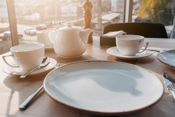 Table setting for breakfast, lunch in sun light. Dinner plate setting with empty plates, cups, teapot near window in cafe, restaurant