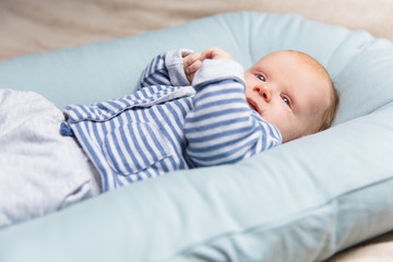 Calm red haired baby in blue and grey clothes, lying on soft pad, looking away. Portrait of cute little child in home interior. Baby care or infant concept