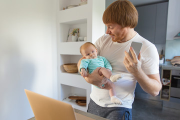 Obraz premium Cheerful new dad working at home, getting good news, sharing with baby daughter. Family portrait of red haired man and cute little child in home interior. Communication or baby care concept
