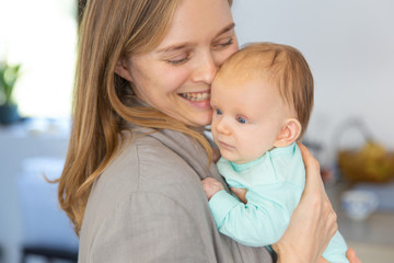 Cheerful mother and positive baby daughter enjoying cuddling. Portrait of young woman and cute little child in home interior. Happy motherhood concept