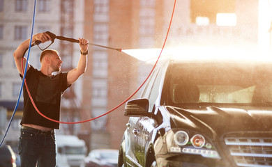 Close-up view of a man cleaning his black car outdoors, water is splashing over the top of the car shining on the sun.