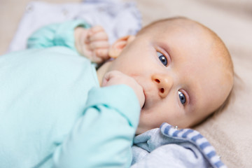 Closeup of pensive sweet baby with finger in mouth lying on bed cover, looking away. Portrait of cute little child in home interior. Childhood or baby care concept