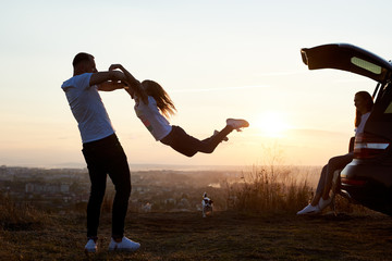 Silhouette of a father swinging his daughter by the arms, mother sitting in the car trunk on top of...