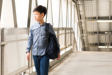 Confident, independent & stylish Asian teenage boy walk in walkway to platform in modern train terminal, look away, carry backpack, commute to school in city as his daily routine in the morning. 