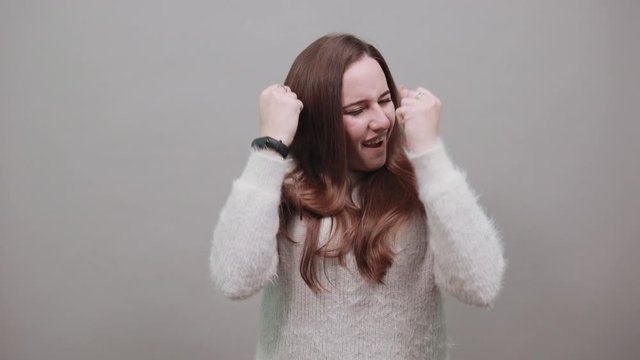 Caucasian attractive woman in sweater keeping fists up, dancing, ready for fight isolated on gray background in studio. People emotions, lifestyle concept.