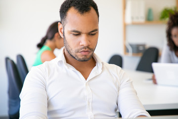 Obraz premium Pensive office worker sitting at table and looking at laptop. Front view of concentrated African American man working in office. Employment concept