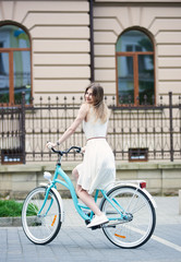 Obraz na płótnie Canvas Cute woman rides the old city streets on a vintage blue bicycle. A girl in a light summer dress with a smile looks at the camera against the background of a building with beautiful architecture