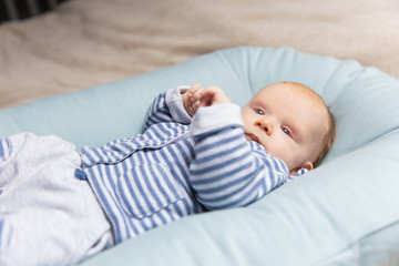 Curious pensive red haired baby in blue and grey clothes, lying on soft mattress, looking away. Portrait of cute little child in home interior. Baby care or childhood concept